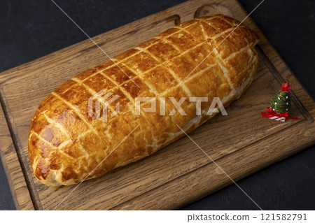 Whole beef Wellington on a wooden board. A small toy Christmas tree nearby. Traditional festive dish of beef tenderloin with mushrooms and Parma ham baked in puff pastry. Selective focus, close-up. 121582791