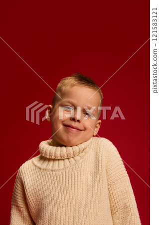 Boy smiles warmly, wearing knitted sweater, tilting head slightly, expressing confidence against deep red background. Boy smiles warmly, wearing knitted sweater, tilting head slightly, expressing confidence against deep red background. 121583121