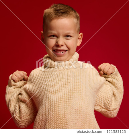 Portrait of smiling boy in beige sweater, holding arms bent, flexing muscles with confident grin against rich red background. Portrait of smiling boy in beige sweater, holding arms bent, flexing muscles with confident grin against rich red background. 121583123