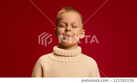 Boy in beige sweater, eyes closed, smiling with peaceful, satisfied expression, standing confidently against rich red background. Boy in beige sweater, eyes closed, smiling with peaceful, satisfied expression, standing confidently against rich red background. 121583138