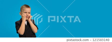 Banner. Young boy in black blazer, white shirt, cupping face with hands, smiling shyly while standing at side against bright blue background. 121583140