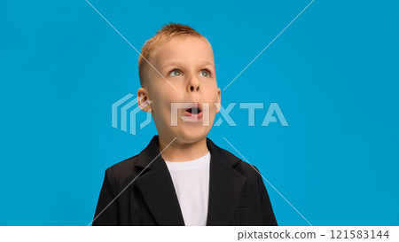 Surprised boy in black blazer and white shirt, wide-eyed, making an exaggerated expression of amazement, radiating curiosity against bright blue background. 121583144