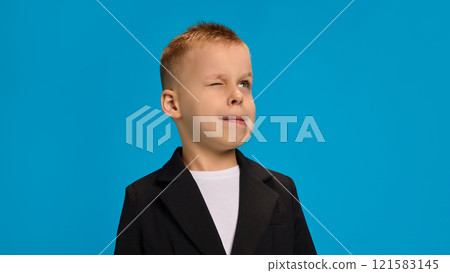 Playful boy in black blazer and white shirt, winking with lips pursed slightly, showcasing a cheeky, fun attitude against bright blue background. Playful boy in black blazer and white shirt, winking with lips pursed slightly, showcasing a cheeky, fun attitude against bright blue background. 121583145