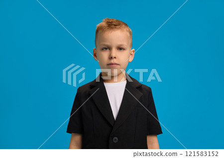 Serious boy dressed in black blazer and white shirt, standing upright, looking directly at camera with calm expression against bright blue background. 121583152