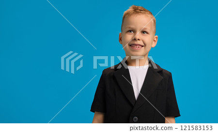 Smiling boy wearing black blazer and white shirt, gazing upward with joyful expression, standing confidently against bright blue background. Smiling boy wearing black blazer and white shirt, gazing upward with joyful expression, standing confidently against bright blue background. 121583153