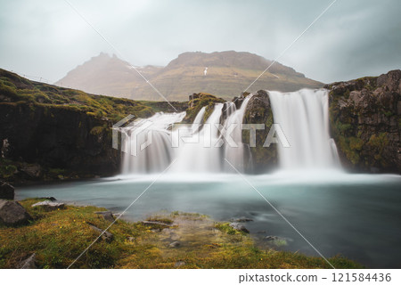 View to the Kirkjufell waterfall at summer. Kirkjufellsfoss waterfall, Iceland, Europe. View to the Kirkjufell waterfall at summer. Kirkjufellsfoss waterfall, Iceland, Europe. 121584436