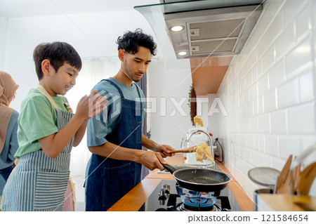 Side view young Asian father show skill of cooking by throwing pan cake up and his son cheer up beside in the kitchen of their house. Side view young Asian father show skill of cooking by throwing pan cake up and his son cheer up beside in the kitchen of their house. 121584654