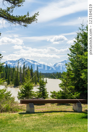 Jasper National Park summer landscape. Wooden bench on Athabasca River bank. Alberta, Canada. Canadian Rockies. Mount Kerkeslin and Mount Hardisty in the background. Jasper National Park summer landscape. Wooden bench on Athabasca River bank. Alberta, Canada. Canadian Rockies. Mount Kerkeslin and Mount Hardisty in the background. 121584819