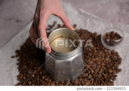 Woman's hand prepares to making coffee in a geyser coffee maker moka machine. Scattered fresh aromatic coffee beans around. Light background Woman's hand prepares to making coffee in a geyser coffee maker moka machine. Scattered fresh aromatic coffee beans around. Light background 121584915