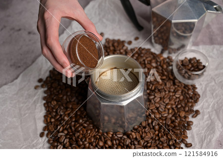 Woman's hands pouring coffee from a small transparent cup in geyser coffee maker moka machine. Scattered fresh aromatic coffee beans around. Light background. Preparation to making coffee 121584916