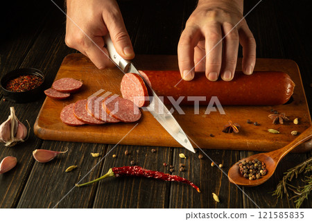 Hands skillfully cut through a rich sausage on a wooden board, surrounded by spices, garlic, and vibrant herbs. The warm atmosphere exudes culinary tradition and passion for food Hands skillfully cut through a rich sausage on a wooden board, surrounded by spices, garlic, and vibrant herbs. The warm atmosphere exudes culinary tradition and passion for food 121585835