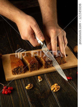 Hands expertly cut into a beautifully baked cake made with nuts and dried fruits. The rustic wooden board contrasts with the vibrant garnishes, creating a warm and inviting culinary moment 121585836