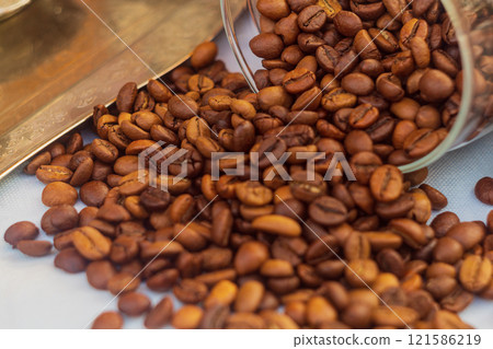 A pile of red roasted coffee beans spilled out of an overturned transparent glass jar onto the white surface of the tablecloth on table. One of the most important export products in some countries 121586219