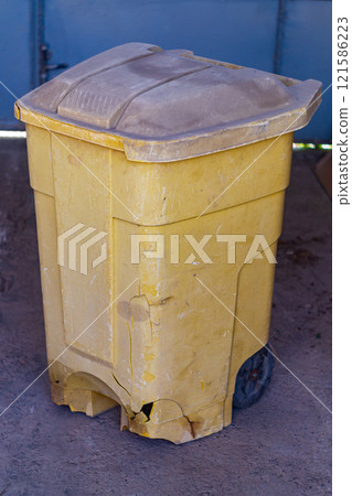 Old orange plastic container for collecting household garbage with black wheels has suffered numerous damage to body in the form of cracks, scratches, chips, scuffs during operation. High angle view 121586223