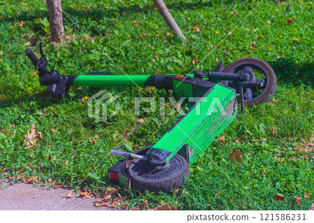 Green electric scooter provided for rent lies on green grass of park. Act of vandalism, improper driving, non-working transport, negligent attitude to rented equipment by user, client, repair costs 121586231