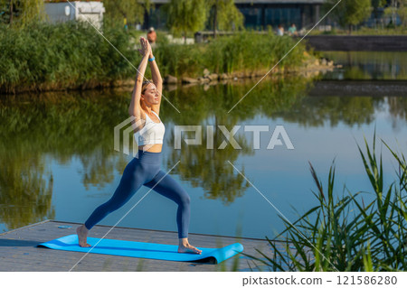 Caucasian woman standing in lunge fitness exercise sport health balance position near pond in park 121586280