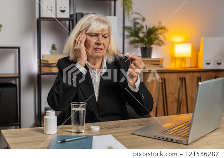 Sick senior businesswoman suffering from headache taking medicine and working on laptop at home Sick senior businesswoman suffering from headache taking medicine and working on laptop at home 121586287