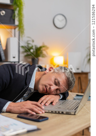 Bored Caucasian young businessman sitting with laptop computer while sleeping at home office desk 121586294