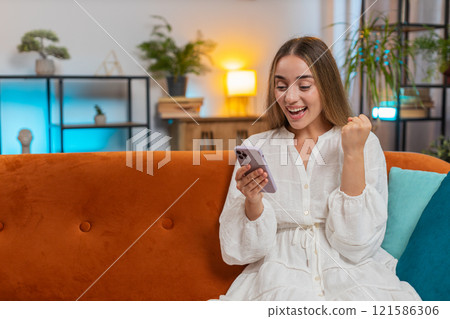 Excited woman sitting on sofa using smartphone typing browsing reading good news celebrating victory Excited woman sitting on sofa using smartphone typing browsing reading good news celebrating victory 121586306