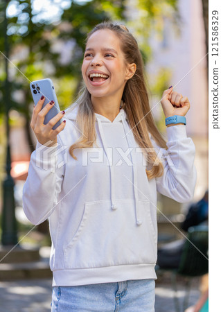 Happy Caucasian young woman using smartphone celebrating win good news, lottery on city town street 121586329