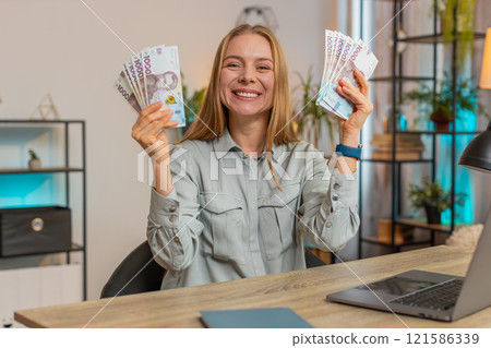 Excited businesswoman freelancer holding fan of Ukrainian hryvnias UAH cash dancing at home office Excited businesswoman freelancer holding fan of Ukrainian hryvnias UAH cash dancing at home office 121586339