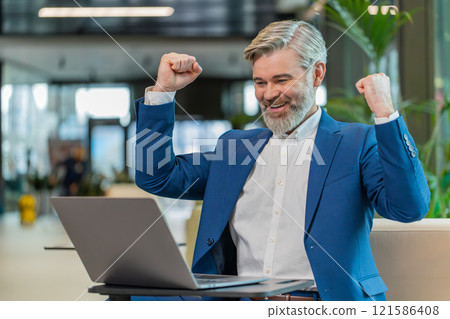 Caucasian bearded businessman working on laptop surprised by sudden victory in modern hotel lobby 121586408