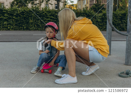 Mom helping her daughter to put on a helmet and ride a skateboard 121586972