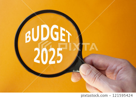 Budget 2025 symbol. Magnifying Glass with words Budget 2024. Beautiful orange background. Businessman hand. Business and Budget 2025 concept. Copy space. Budget 2025 symbol. Magnifying Glass with words Budget 2024. Beautiful orange background. Businessman hand. Business and Budget 2025 concept. Copy space. 121595424