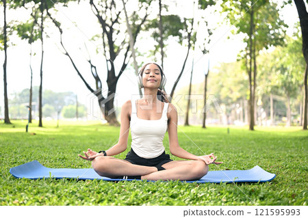 Calm young woman wearing headphones practicing meditation on a grassy lawn in a serene park. 121595993