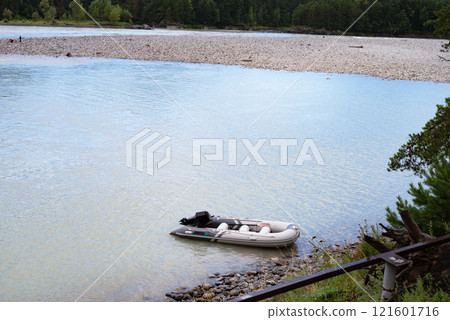 Inflatable boat moored on a serene riverbank with forest backdrop. Inflatable boat moored on a serene riverbank with forest backdrop. 121601716