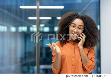 Portrait of a joyful businesswoman, office worker with a phone in her hands talking to clients, smiling and looking at the camera, standing near the window of an office building at her workplace. Portrait of a joyful businesswoman, office worker with a phone in her hands talking to clients, smiling and looking at the camera, standing near the window of an office building at her workplace. 121603960