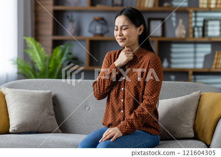 Asian woman at home sits on couch feeling throat discomfort. Expression of concern suggests health issue or pain. Relaxed home setting, expressing pain or worry in her expression. Asian woman at home sits on couch feeling throat discomfort. Expression of concern suggests health issue or pain. Relaxed home setting, expressing pain or worry in her expression. 121604305