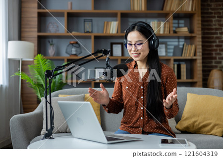 Asian woman hosting a podcast at home, speaking into a microphone while using a laptop. Wearing headphones, she appears engaged and lively, surrounded by a comfortable setting. Smartphone visible. 121604319