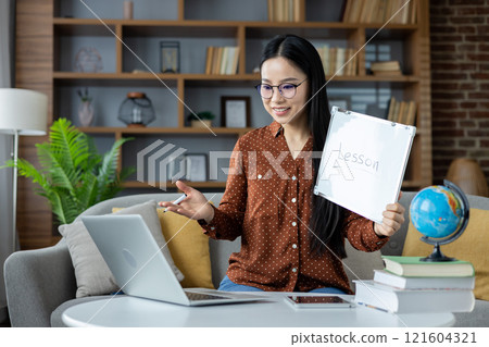 Asian woman conducting online lesson from home. She sits on sofa with laptop, holding a whiteboard marked Lesson, engaged in interactive teaching session, conveying education and technology. Asian woman conducting online lesson from home. She sits on sofa with laptop, holding a whiteboard marked Lesson, engaged in interactive teaching session, conveying education and technology. 121604321