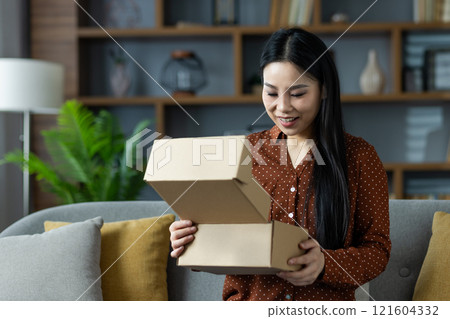 Asian woman excitedly opens package box on sofa in home setting. Scene captures joy and anticipation of unboxing. Woman's expression of delight signifies receiving gift or purchase. Asian woman excitedly opens package box on sofa in home setting. Scene captures joy and anticipation of unboxing. Woman's expression of delight signifies receiving gift or purchase. 121604332
