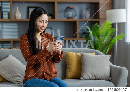 Young Asian woman in cozy home setting uses smartphone, seated comfortably on sofa. She engages in online communication, expressing happiness and relaxation in domestic environment. Young Asian woman in cozy home setting uses smartphone, seated comfortably on sofa. She engages in online communication, expressing happiness and relaxation in domestic environment. 121604437