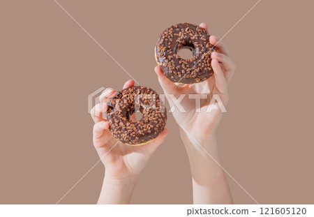 Hands holding two glazed chocolate donuts on mocha brown background. 121605120
