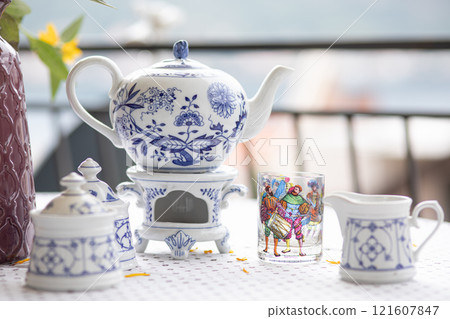 Elegant tea set displayed on a table featuring a decorative teapot, teacup, and creamer, showcasing intricate blue floral patterns and a colorful glass, highlighting a serene afternoon setting Elegant tea set displayed on a table featuring a decorative teapot, teacup, and creamer, showcasing intricate blue floral patterns and a colorful glass, highlighting a serene afternoon setting 121607847