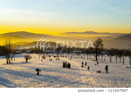 The sun sets behind the snow-covered hills of Prichovice, casting a warm glow over skiers and families enjoying the winter wonderland. The serene landscape captures the beauty of winter. 121607907
