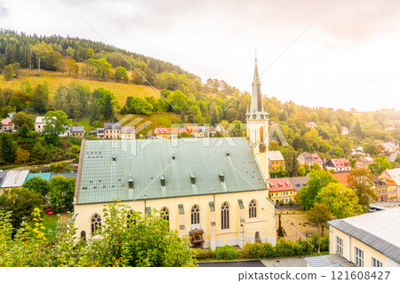 The Church of St. Joachim stands proudly in Jachymov, surrounded by lush greenery and colorful houses. Sunlight breaks through clouds, illuminating this serene setting. 121608427