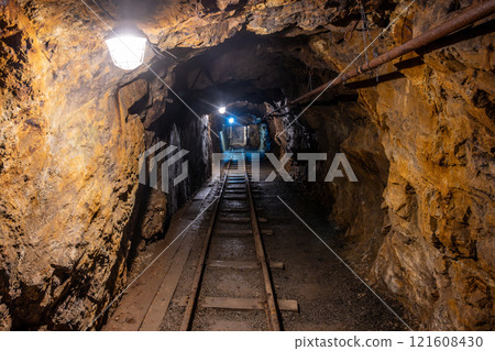 Deep within the Jachymov mine shaft in Czechia, a narrow tunnel reveals old mining tracks and ambient lights illuminating the rocky walls. This location holds a rich history of mineral extraction. 121608430