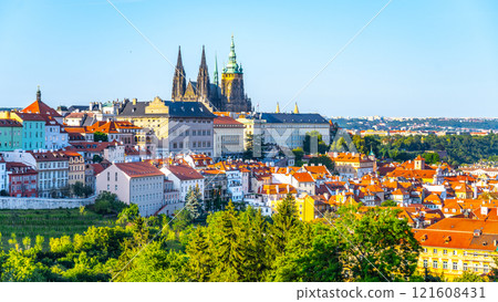 The grand architecture of Prague Castle towers above the vibrant rooftops of the city. Lush greenery complements the lively urban landscape, creating a picturesque view on a clear day. 121608431