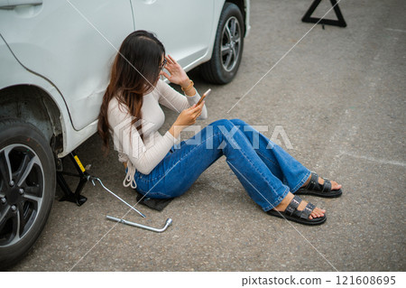 A woman dressed in jeans is struggling with car trouble while using her phone urgently 121608695