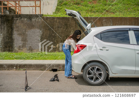 A woman is actively fixing car troubles on the roadside while dressed in a casual outfit A woman is actively fixing car troubles on the roadside while dressed in a casual outfit 121608697
