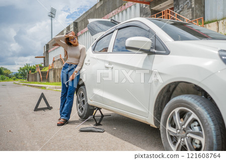 A woman is diligently repairing a car tire on a rural road while using safety equipment 121608764