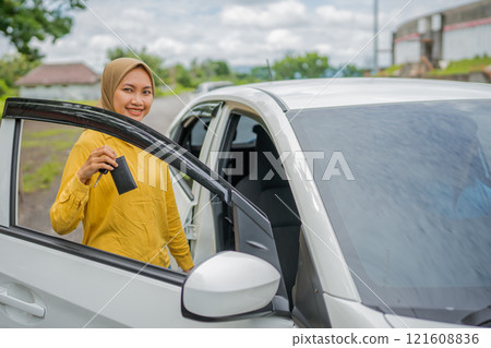 A smiling woman confidently holds a car key next to her white vehicle in daylight 121608836