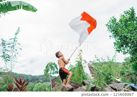 unclothed child raises the Indonesian flag excitedly with nature in the background 121608867