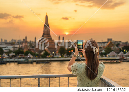 tourist woman enjoys view to Wat Arun Temple in sunset, Traveler take photo to Temple of Dawn by smartphone from rooftop bar. Landmark and Travel destination in Bangkok, Thailand and Southeast Asia 121612832