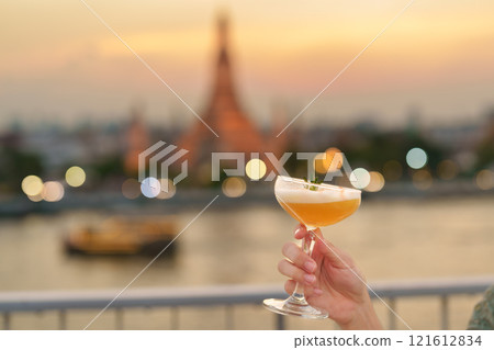 tourist hand shows cocktail glass during enjoy view to Wat Arun Temple in sunset, Traveler visits Temple of Dawn near from rooftop bar. Travel destination in Bangkok, Thailand and Southeast Asia tourist hand shows cocktail glass during enjoy view to Wat Arun Temple in sunset, Traveler visits Temple of Dawn near from rooftop bar. Travel destination in Bangkok, Thailand and Southeast Asia 121612834