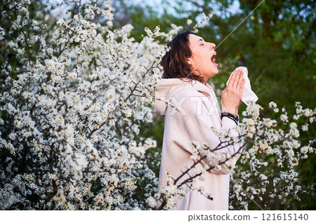 Man allergic suffering from seasonal allergy at spring in blossoming garden at springtime. Young man sneezing and blowing nose with nasal handkerchief in front of blooming tree. Spring allergy concept Man allergic suffering from seasonal allergy at spring in blossoming garden at springtime. Young man sneezing and blowing nose with nasal handkerchief in front of blooming tree. Spring allergy concept 121615140